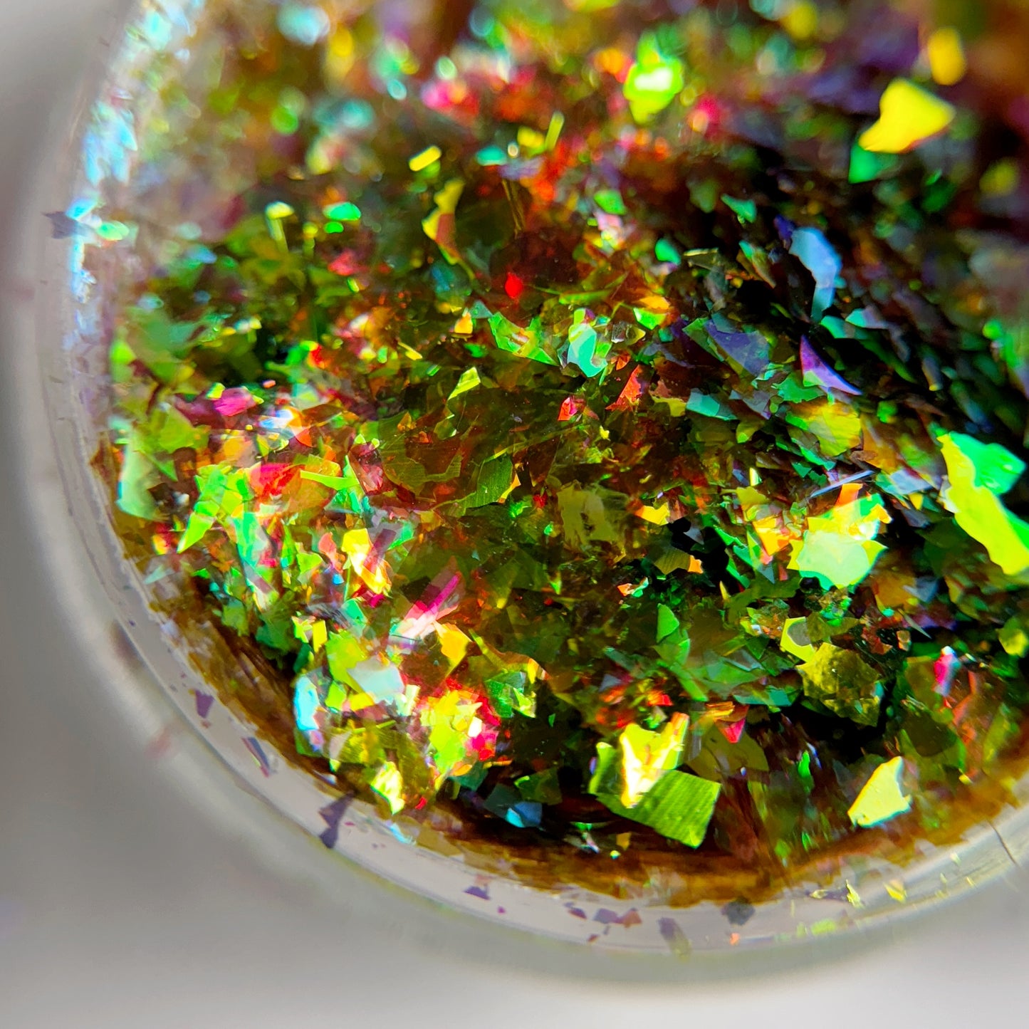 A close-up image of multicolored flaky nail art powder, with a predominance of green, reflecting light and creating a shimmering effect.