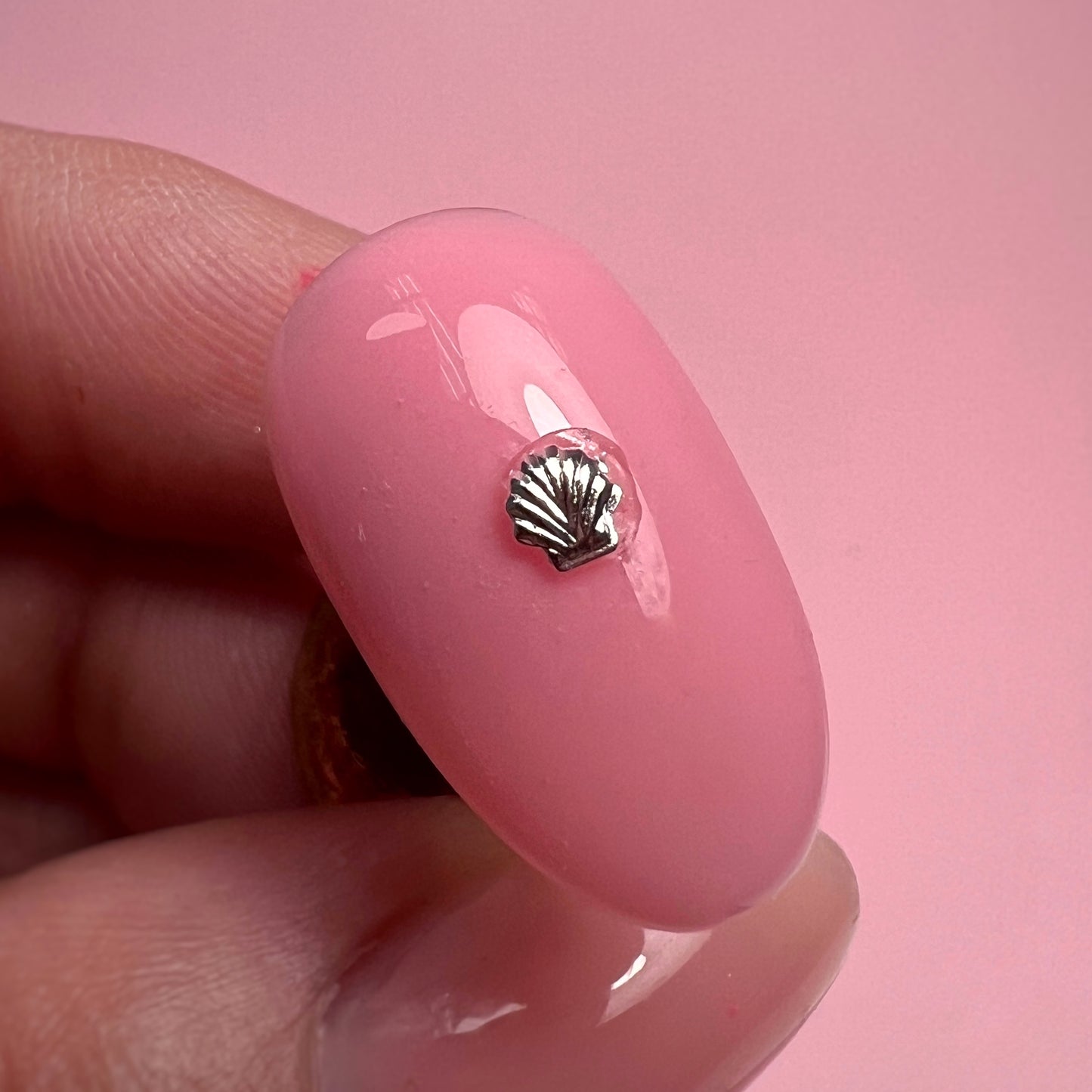 Pink nail with a silver shell charm held by a hand against a pink background