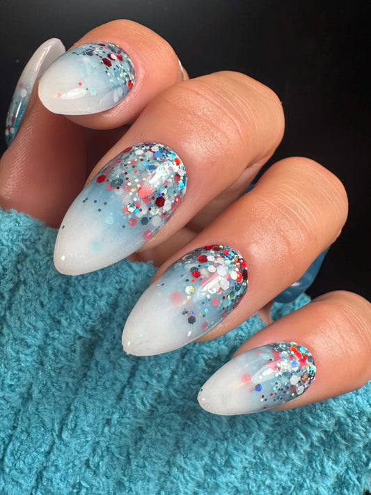 Nails with white base and multicolored glitter design on a blue textured surface.