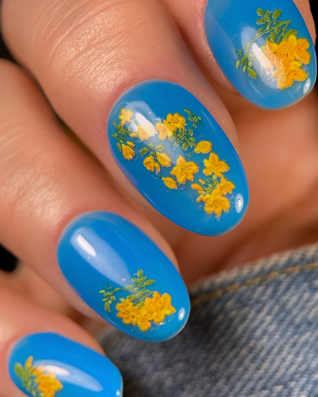 Nails with blue polish and yellow-orange floral design against denim background.