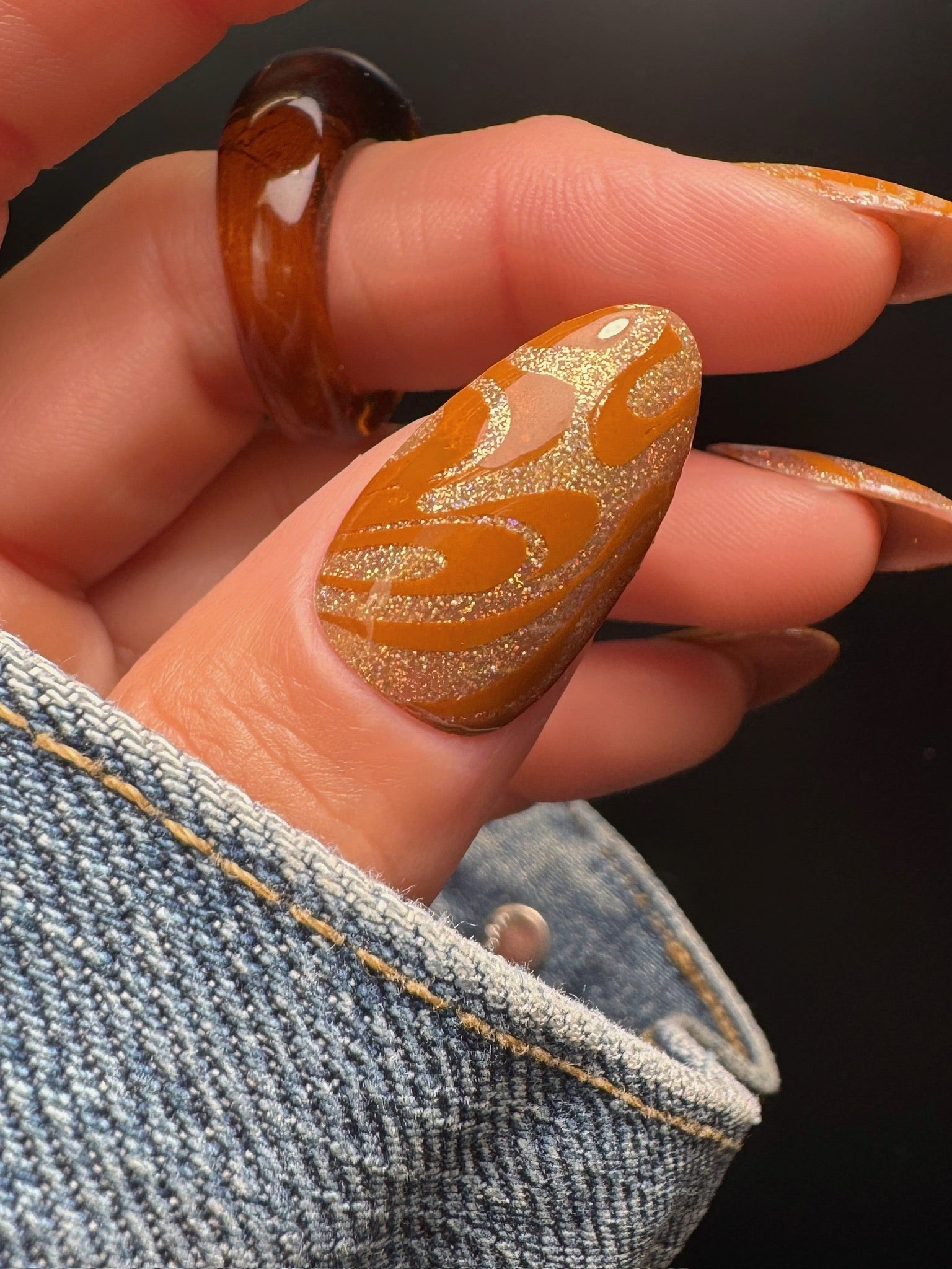 Manicured hand with ring with nails featuring gold and dark yellow  patterns against a dark background
