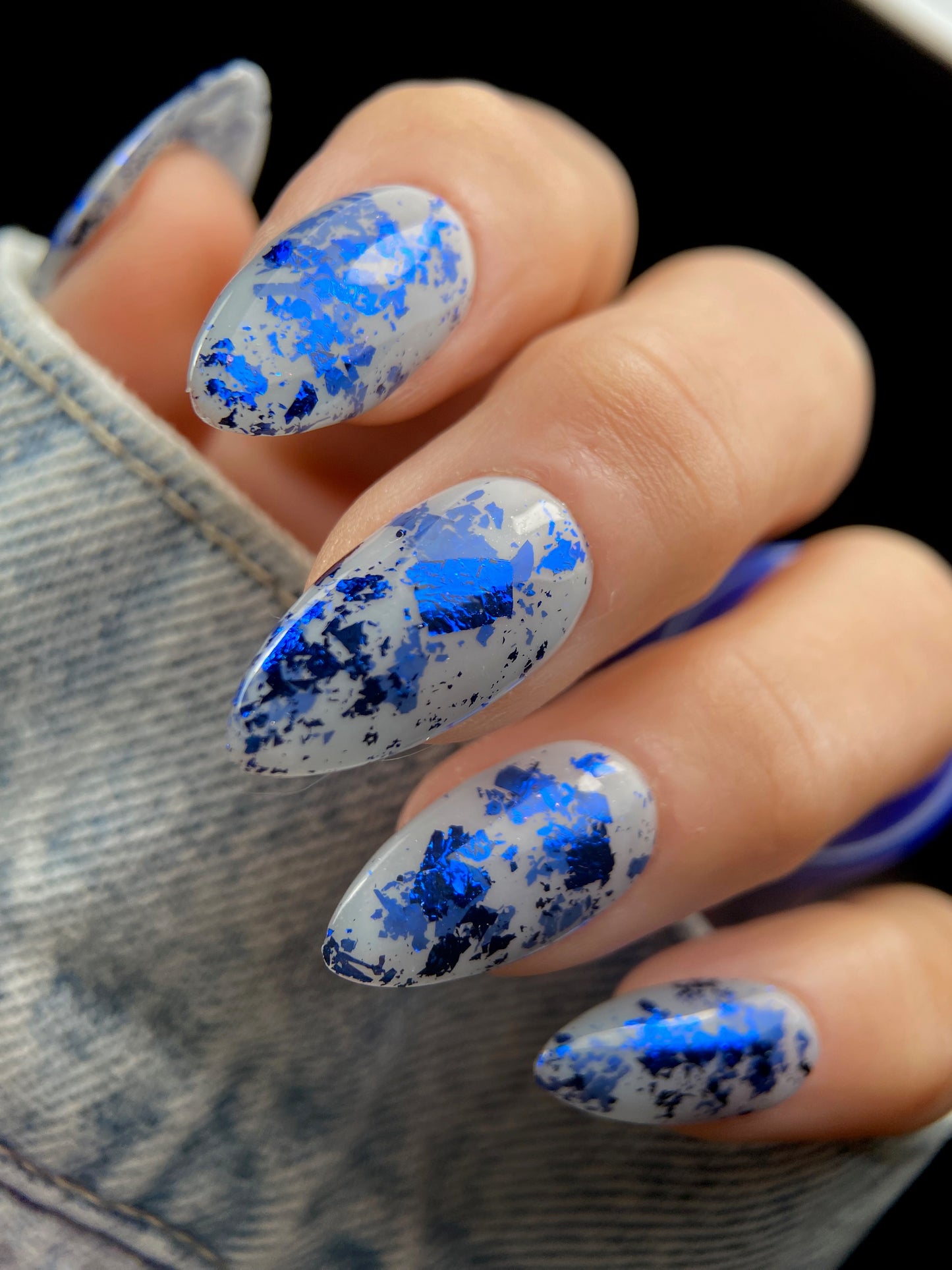 Close-up of almond-shaped nails with navy blue metallic foil flakies scattered over a sheer milky gray base.