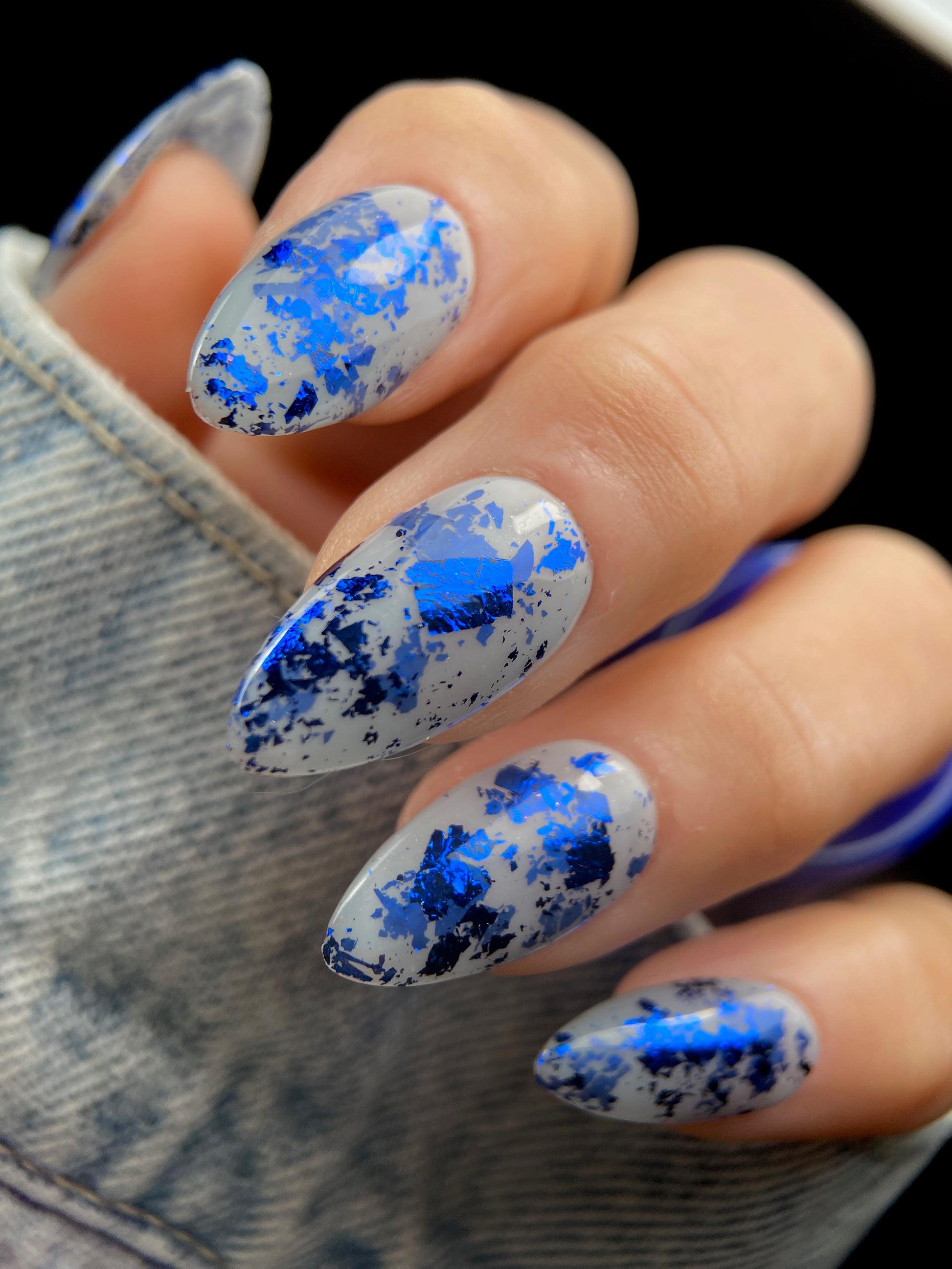 Close-up of almond-shaped nails with navy blue metallic foil flakies scattered over a sheer milky gray base.