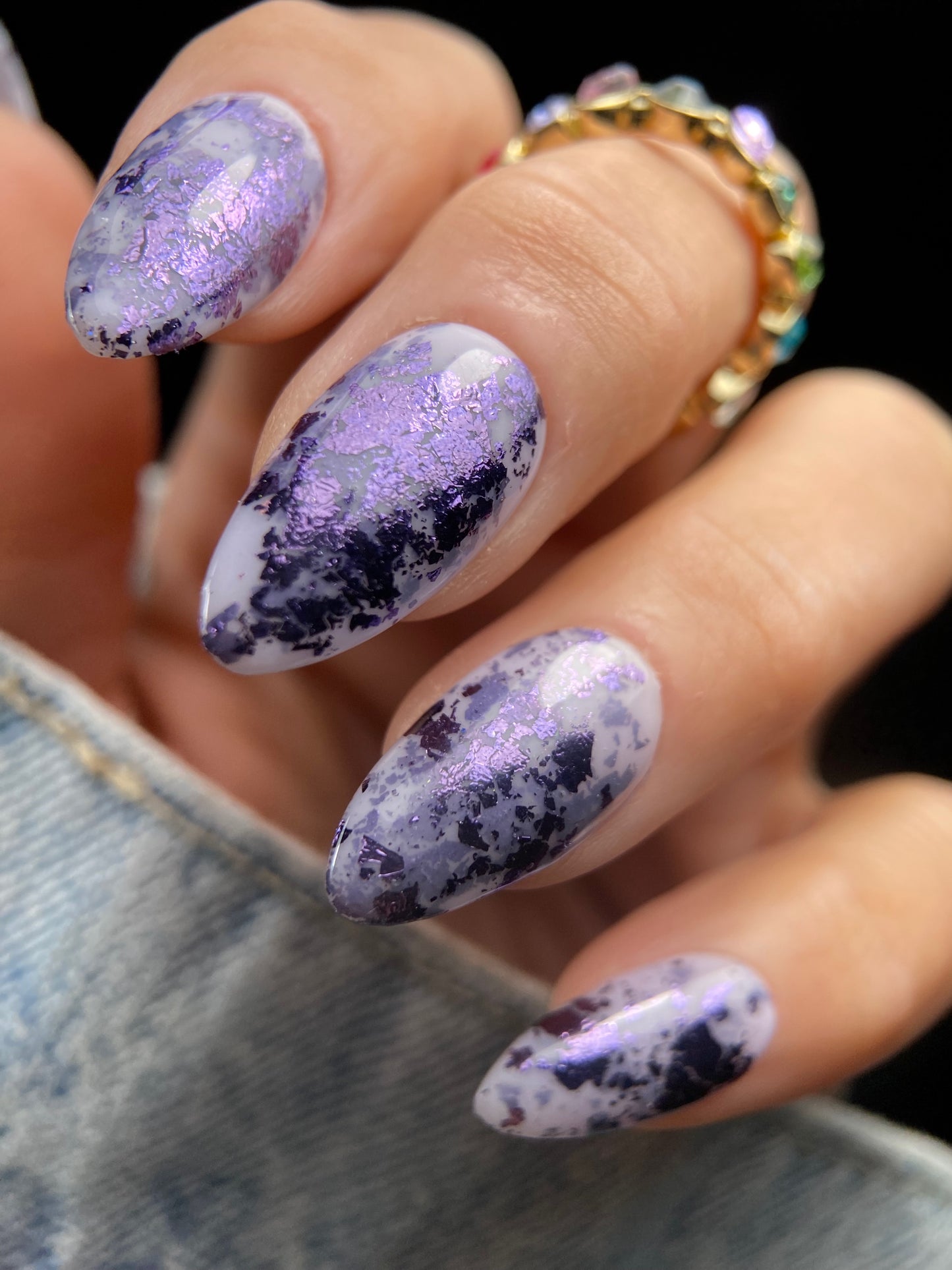 Close-up of almond-shaped nails with plum and lavender metallic foil flakies scattered over a sheer milky lavender base.