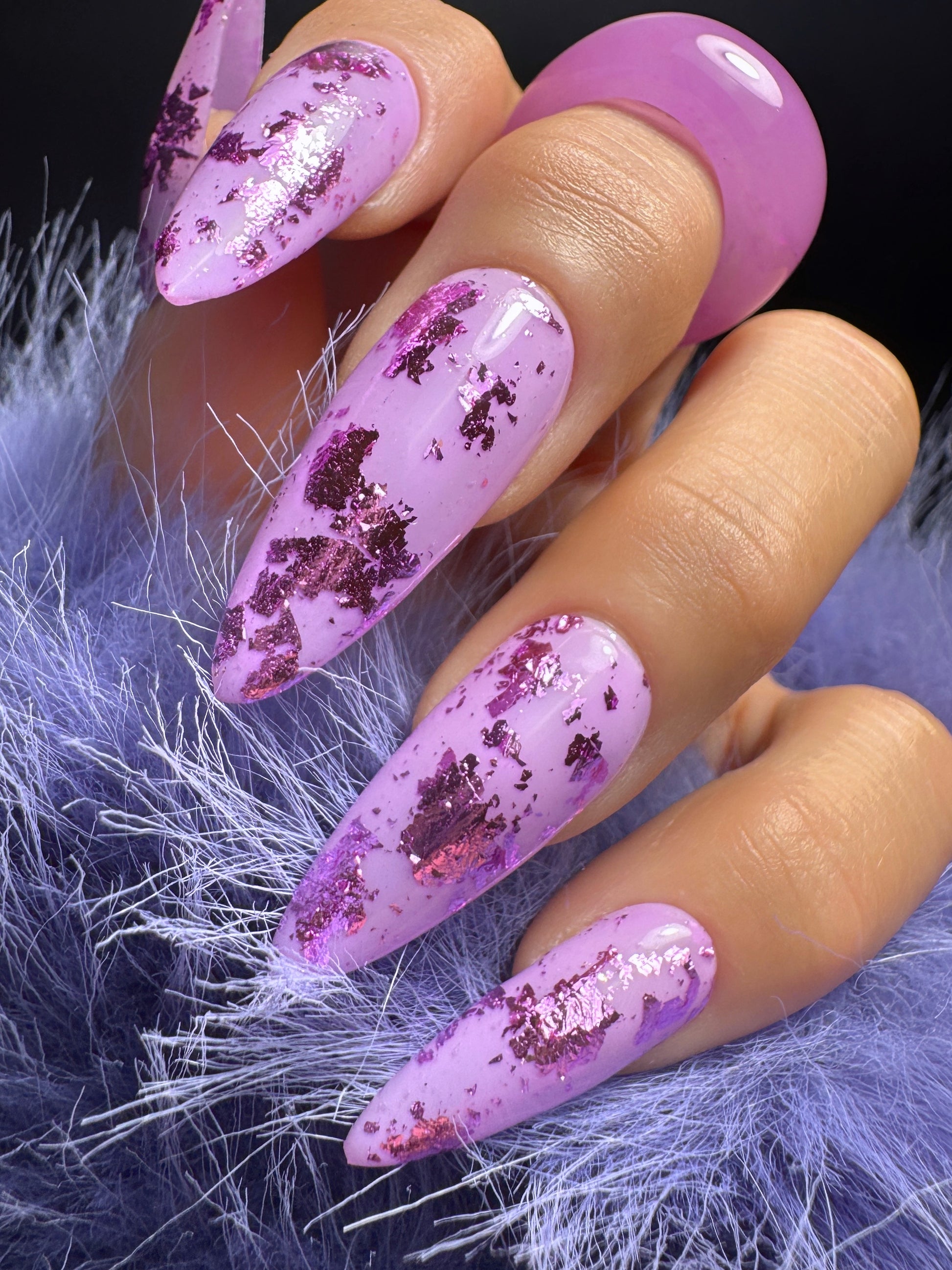Hand with purple acrylic nails featuring purple foil flakes on a textured purple background
