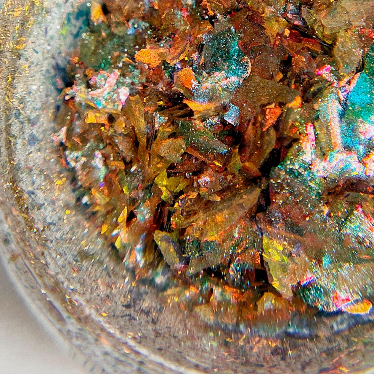 A close-up image of a sparkling, multicolored nail art flake, showing hues of blue, gold, and red.